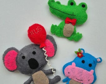 Op de afbeelding: Handgemaakte vilten dierenfiguren, waaronder een groene alligator met een rode strik, een grijze koala met een rode ballon met "HUG" en een blauwe nijlpaard met een bloem. Waarschijnlijk kinderspeelgoed of decoratie-artikelen.