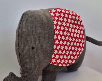 Può includere: Un elefante giocattolo di stoffa grigia con una coperta a fantasia floreale rossa e bianca sulla schiena. L'elefante ha una proboscide, piccole orecchie e una coda. Il giocattolo è in tessuto e ha un design semplice.