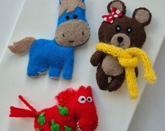 Op de afbeelding: Drie vilten dierenfiguren: een blauw paard, een bruine beer met een gele sjaal en een rode strik, en een rood paard met groene vlekken. De paarden hebben bruine manen en staarten. De beer heeft een gele sjaal.