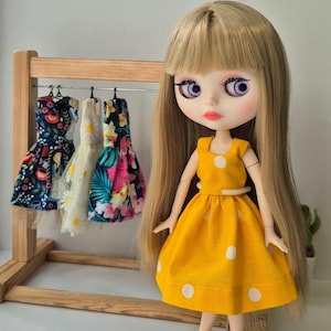 Puede incluir: Una muñeca con pelo largo rubio y ojos morados que lleva un vestido amarillo con lunares blancos. Detrás de la muñeca hay un perchero de madera con tres vestidos en miniatura con estampados florales. La muñeca lleva zapatos marrones y rojos.