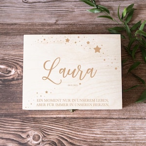 Könnte beinhalten: Eine Holzkiste mit einer goldenen Inschrift, die "Laura" und "Ein Moment nur in unserem Leben, aber fur immer in unseren Herzen" lautet. Die Kiste ist mit einer Streuung von goldenen Sternen verziert.