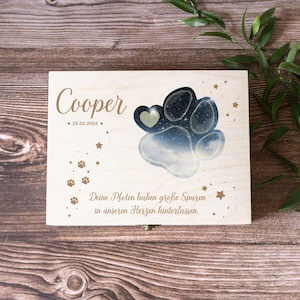Könnte beinhalten: Eine Holzkiste mit einem Pfotenabdruck-Design und dem Text "Cooper * 25.02.2024 *" und "Deine Pfoten haben große Spuren in unseren Herzen hinterlassen." Die Kiste steht auf einer Holzoberfläche.