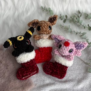 Puede incluir: Tres figuras de peluche de ganchillo en calcetines navideños. Las figuras son un conejito marrón, una criatura negra y amarilla y una criatura rosa. Los calcetines son rojos con ribete blanco. El texto "@mokka_crochet" está en un calcetín.