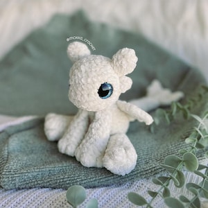 May include: A white crocheted dragon plushie with large blue eyes, small wings, and four legs. The dragon is sitting on a green blanket. The text "@mokka_crochet" is visible in the image.