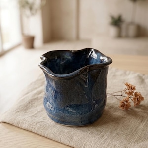 May include: A deep blue ceramic vessel with a glossy, mottled glaze and an irregular, wavy rim. The pot features a rounded body with subtle indentations along the sides, creating a textured appearance. The interior is dark and smooth.