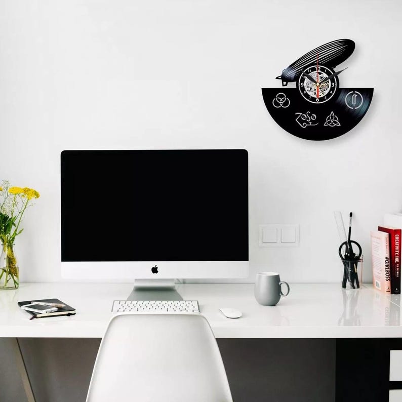 K&ouml;nnte beinhalten: Eine schwarze Vinyl-Schallplattenuhr mit einem Zeppelin-Luftschiff-Design. Das Zifferblatt zeigt traditionelle Zahlen und Zeiger. Die Uhr ist an einer wei&szlig;en Wand &uuml;ber einem Schreibtisch mit Computer und Zubeh&ouml;r angebracht.