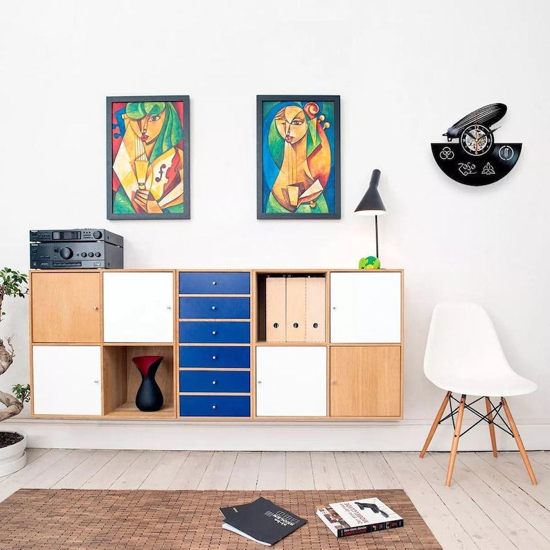K&ouml;nnte beinhalten: Ein modernes Wohnzimmer mit einem an der Wand befestigten Schrank mit Holz-, wei&szlig;en und blauen Stauf&auml;chern. Zwei gerahmte abstrakte Kunstwerke h&auml;ngen &uuml;ber dem Schrank. Eine schwarze Vinyl-Schallplattenuhr und ein wei&szlig;er Stuhl vervollst&auml;ndigen die Einrichtung.