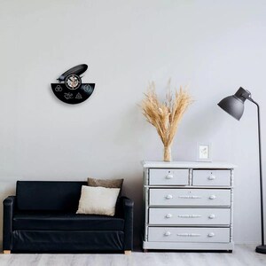K&ouml;nnte beinhalten: Eine schwarze Vinyl-Schallplattenuhr mit einem Zeppelin-Design und verschiedenen Symbolen. Die Uhr ist an einer wei&szlig;en Wand befestigt, mit einem schwarzen Sofa, einer grauen Kommode und einer Stehlampe im Hintergrund. Die Zeiger der Uhr zeigen die Zeit an.