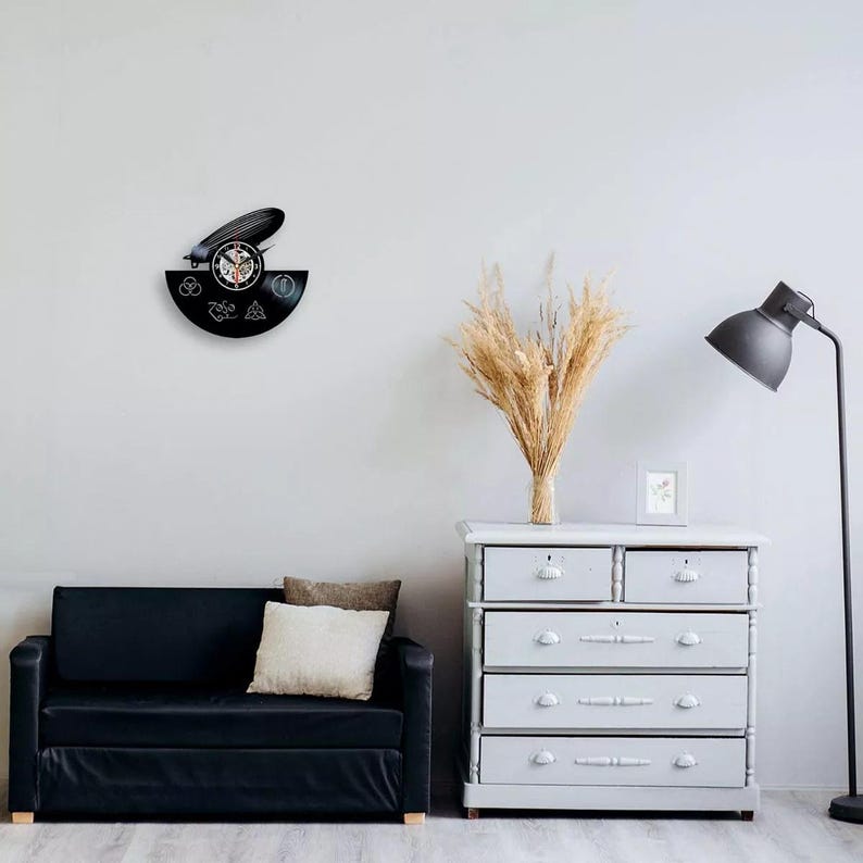 K&ouml;nnte beinhalten: Eine schwarze Vinyl-Schallplattenuhr mit einem Zeppelin-Design und verschiedenen Symbolen. Die Uhr ist an einer wei&szlig;en Wand befestigt, mit einem schwarzen Sofa, einer grauen Kommode und einer Stehlampe im Hintergrund. Die Zeiger der Uhr zeigen die Zeit an.