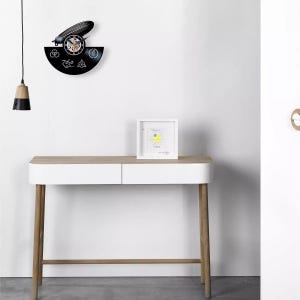 K&ouml;nnte beinhalten: Eine schwarze Vinyl-Schallplattenuhr mit Zeppelin-Design und Zifferblatt. Die Uhr ist an einer wei&szlig;en Wand &uuml;ber einem wei&szlig;en und holzfarbenen Konsolentisch angebracht. Ein gerahmtes Bild und eine H&auml;ngeleuchte sind ebenfalls zu sehen.