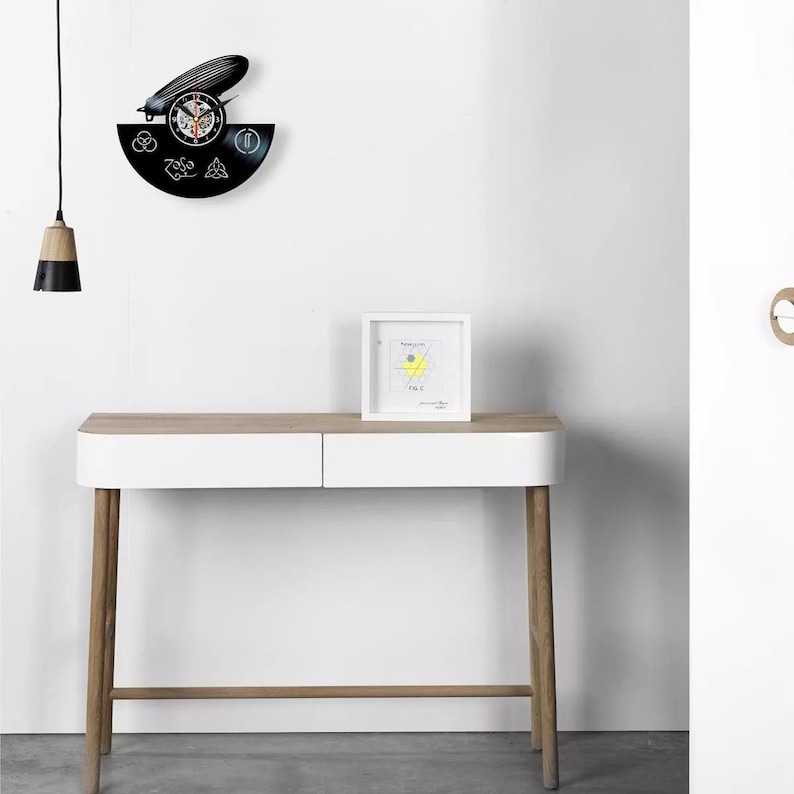K&ouml;nnte beinhalten: Eine schwarze Vinyl-Schallplattenuhr mit Zeppelin-Design und Zifferblatt. Die Uhr ist an einer wei&szlig;en Wand &uuml;ber einem wei&szlig;en und holzfarbenen Konsolentisch angebracht. Ein gerahmtes Bild und eine H&auml;ngeleuchte sind ebenfalls zu sehen.