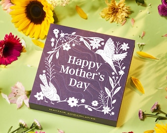 Può includere: Scatola regalo viola con un designo floreale bianco e il testo "Happy Mother's Day". La scatola è circondata da fiori.