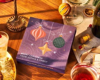 Può includere: Scatola regalo viola con un design natalizio con ornamenti e una stella. La scatola ha il testo "Merry Christmas from Sophia & Family" e "Mayfair Chocolatier" stampato su di essa.