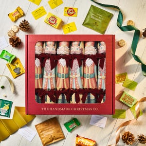 Op de afbeelding: Een rode doos met kerstcrackers met kerstmanontwerpen, omringd door theezakjes, snoepjes en spelkaarten. Op de doos staat "THE HANDMADE CHRISTMAS CO."