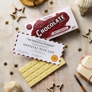 Op de afbeelding: Een witte chocoladereep en een doos witte chocolade van The North Pole Chocolate Company worden feestelijk gepresenteerd. Op de doos staat "White Chocolate" en "Original Est 1892". Een "Official Nice List" certificaat is ook aanwezig.