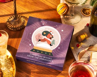 Può includere: Una scatola viola con un'illustrazione di palla di neve che rappresenta Babbo Natale che porta un sacco di regali. La scatola ha il testo "Christmas Wishes from the Jamiesons' Mayfair Chocolatier" stampato su di essa.