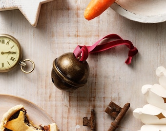 May include: A festive still life featuring a bronze bell with a red ribbon, a pocket watch, a star-shaped ornament, carrots, a mince pie, and antique keys. The items are arranged on a light-colored wooden surface, evoking a holiday theme.