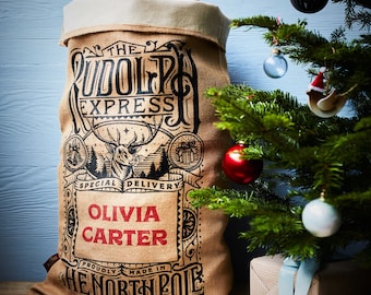 May include: A brown burlap Santa sack with the text "The Rudolph Express Special Delivery" and "Proudly Made in The North Pole". The sack has a red and white name tag with the name "Olivia Carter" printed on it.