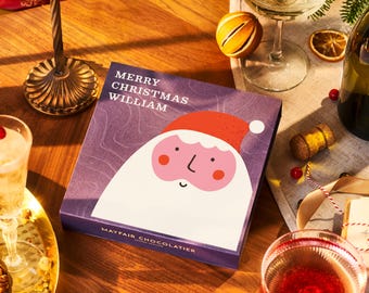 Può includere: Una scatola viola con un'illustrazione di Babbo Natale e il testo "MERRY CHRISTMAS WILLIAM" su un tavolo di legno. La scena comprende calici di champagne, fette d'arancia essiccate e una bottiglia di champagne.