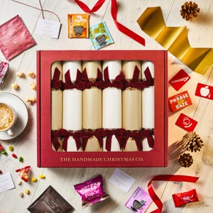 May include: A festive Christmas gift set featuring a red box with cream and beige crackers tied with red velvet bows. The box is surrounded by tea packets, popcorn, candies, and decorative ribbons. The text "THE HANDMADE CHRISTMAS CO." is printed on the box.