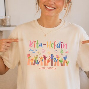 May include: A cream-colored t-shirt featuring the text Kita-Heldin in colorful letters, accompanied by a graphic of various multi-colored handprints and small icons like hearts, a sun, and a tree, with the name Johanna printed below in pink.
