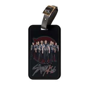 May include: Black luggage tag with a leather strap and gold buckle. The tag features a group of people in black outfits against a red background. The words "Stray Kids" are written in a stylized font.