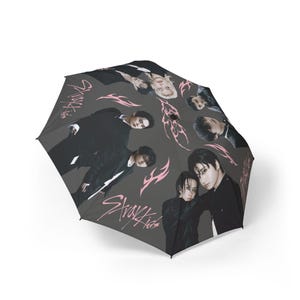 Puede incluir: Un paraguas gris oscuro con un patrón repetido de personas con trajes negros y diseños tribales rosas. El paraguas también tiene el texto "Stray Kids" en rosa.