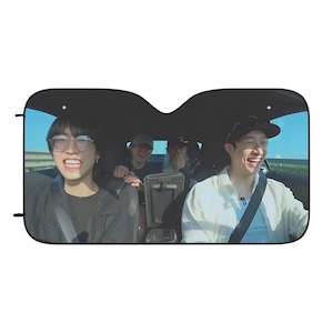 May include: Four people are riding in a car. The person in the front passenger seat is wearing a white shirt and smiling. The person in the back seat on the left is wearing a black shirt and glasses. The person in the back seat on the right is wearing a black hat and a white shirt. The car is driving on a road with a blue sky in the background.