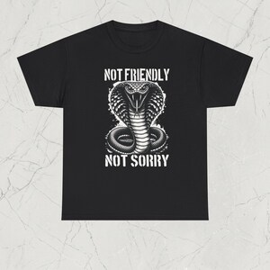 Può includere: T-shirt nera con un'illustrazione in scala di grigi di un cobra. Il serpente è raffigurato con il cappuccio aperto. Il testo "NOT FRIENDLY" e "NOT SORRY" è stampato sopra e sotto il disegno del cobra.