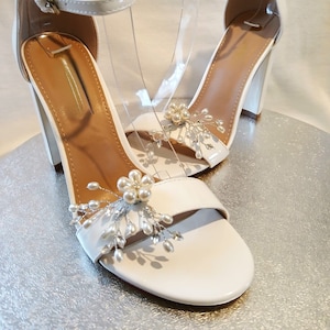May include: White heeled sandals with ankle straps and pearl embellishments. The sandals have a block heel and an open toe.