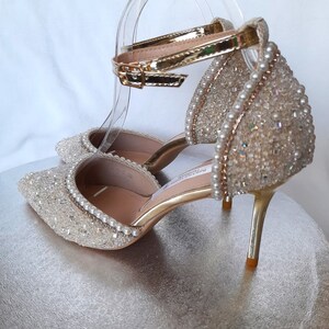 May include: A pair of gold high-heeled shoes with a sparkly, pearl-embellished design. The shoes have an ankle strap with a gold buckle.