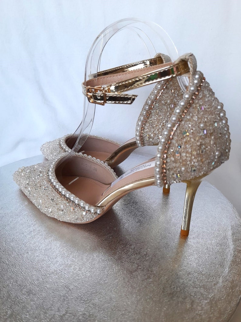 May include: A pair of gold high-heeled shoes with a sparkly, pearl-embellished design. The shoes have an ankle strap with a gold buckle.