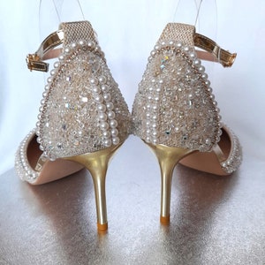 May include: A pair of gold high-heeled shoes with a delicate ankle strap. The shoes are covered in a shimmering fabric adorned with pearls and rhinestones.