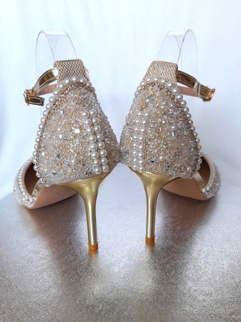 May include: A pair of gold high-heeled shoes with a delicate ankle strap. The shoes are covered in a shimmering fabric adorned with pearls and rhinestones.