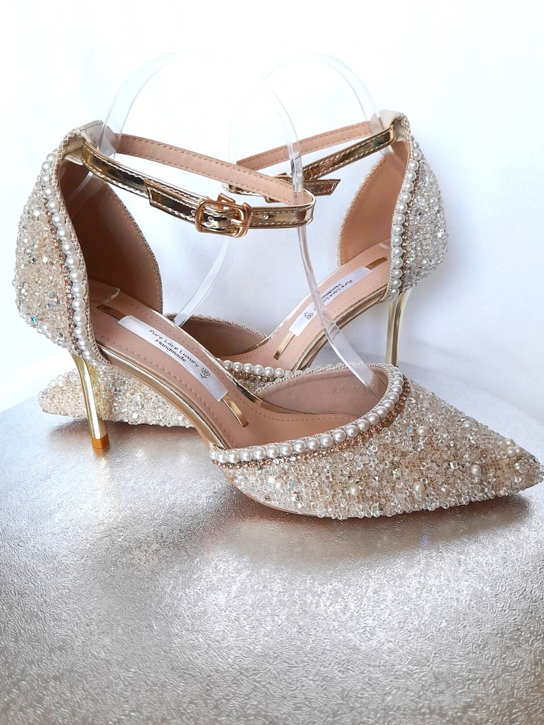 May include: A pair of gold and ivory bridal shoes with a delicate ankle strap and pearl embellishments. The shoes have a stiletto heel and a pointed toe.
