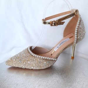 May include: Gold high-heeled shoes with a delicate ankle strap and a pointed toe. The shoes are covered in sparkling crystals and pearls.