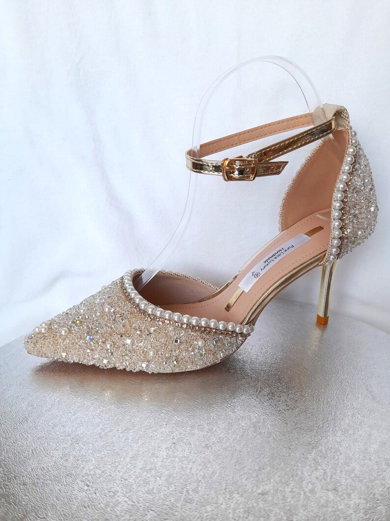 May include: Gold high-heeled shoes with a delicate ankle strap and a pointed toe. The shoes are covered in sparkling crystals and pearls.