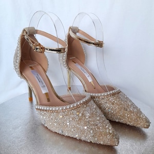 May include: A pair of gold and ivory bridal heels with a delicate ankle strap and a pointed toe. The shoes are embellished with small pearls and rhinestones.