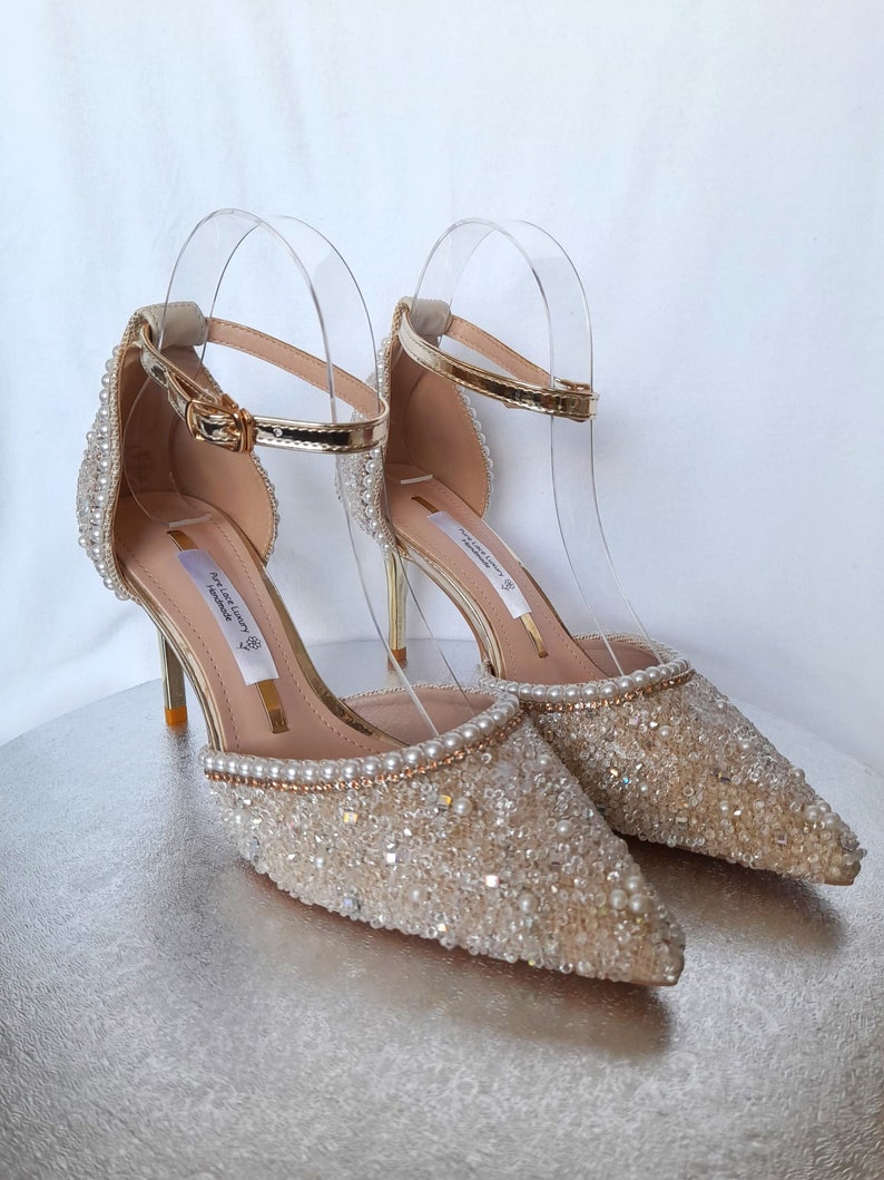 May include: A pair of gold and ivory bridal heels with a delicate ankle strap and a pointed toe. The shoes are embellished with small pearls and rhinestones.