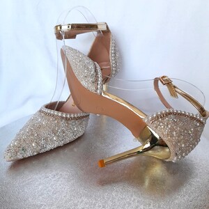 May include: A pair of gold high-heeled shoes with a sparkly, beaded design. The shoes have a pointed toe and a delicate ankle strap.