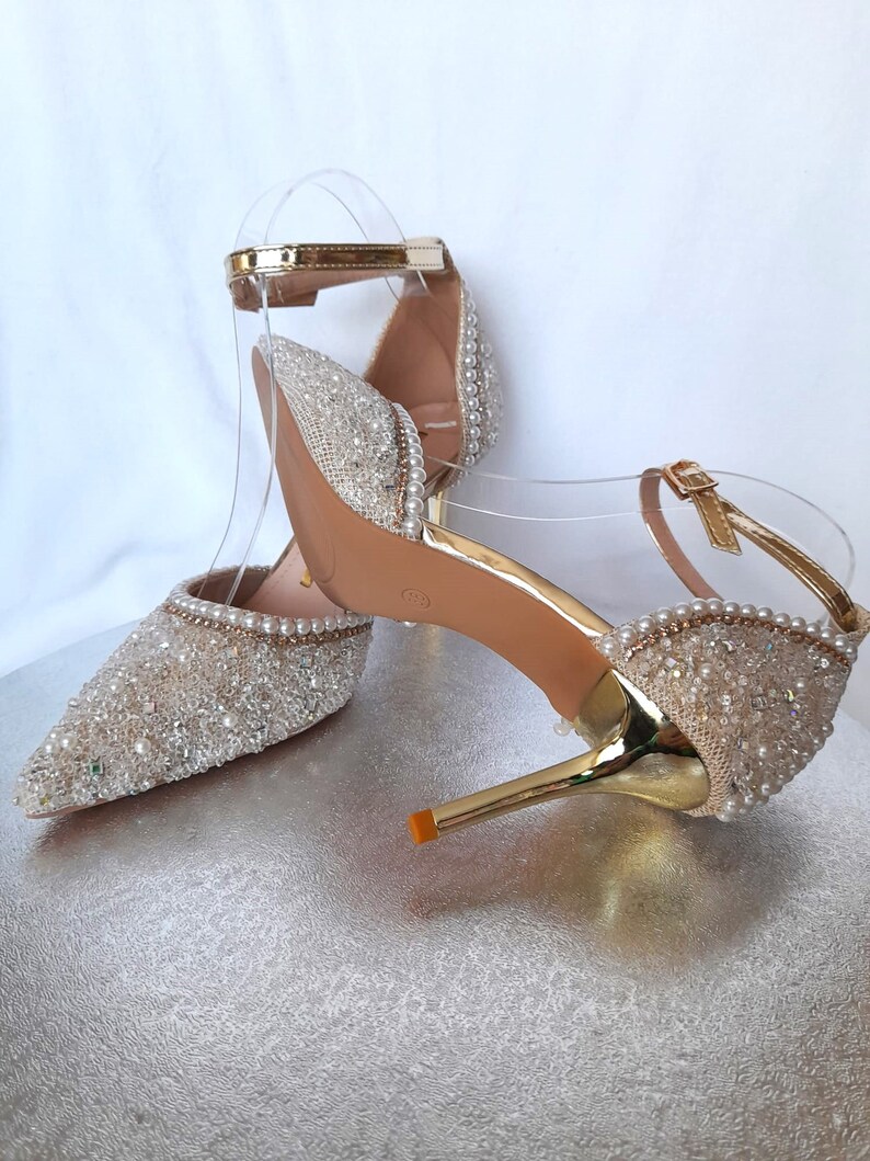 May include: A pair of gold high-heeled shoes with a sparkly, beaded design. The shoes have a pointed toe and a delicate ankle strap.