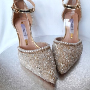 May include: A pair of gold and ivory bridal shoes with a pointed toe and ankle strap. The shoes are covered in small pearls and rhinestones. The text "The Luxury Handmade" is visible on the inside of the shoe.