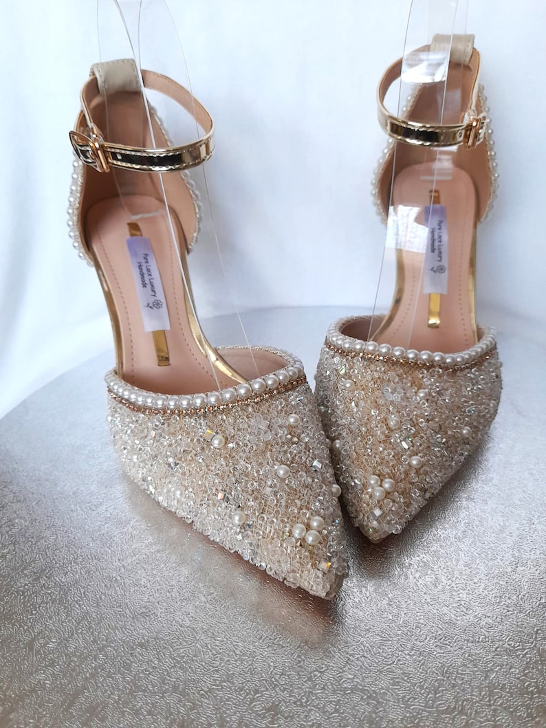 May include: A pair of gold and ivory bridal shoes with a pointed toe and ankle strap. The shoes are covered in small pearls and rhinestones. The text "The Luxury Handmade" is visible on the inside of the shoe.