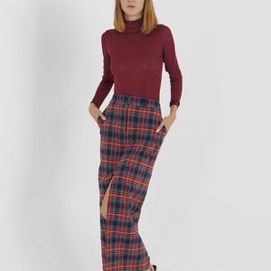Puede incluir: Una mujer con un jersey de cuello alto color burdeos y una falda de cuadros rojos, azules y negros con una abertura delantera. Lleva sandalias de tac&oacute;n negras.