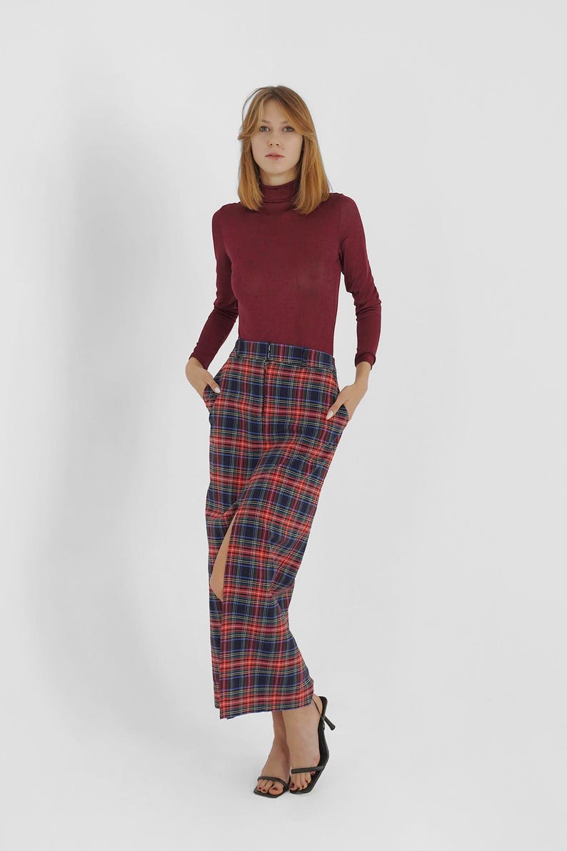 Puede incluir: Una mujer con un jersey de cuello alto color burdeos y una falda de cuadros rojos, azules y negros con una abertura delantera. Lleva sandalias de tac&oacute;n negras.