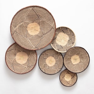 May include: A collection of seven woven baskets in varying sizes. The baskets feature intricate geometric patterns in black and tan, with a circular design in the center. The rims are finished with a dark brown border.