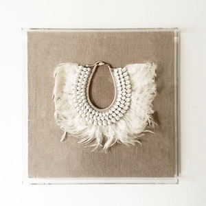 May include: A white feather and shell necklace displayed in a clear acrylic frame on a beige linen background.