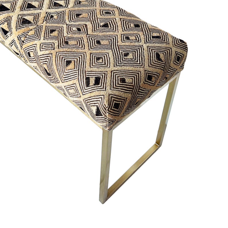 May include: A bench with a geometric patterned seat in beige, black, and brown tones. The bench has a rectangular seat and gold-colored metal legs. The legs are a simple, modern design.