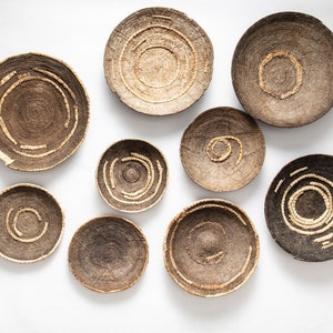 Pode incluir: Nove cestas redondas de vime com padrões intrincados em tons de marrom e bege, penduradas em uma parede branca.