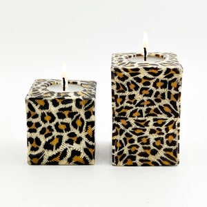 May include: Two square candle holders with a leopard print design. The candle holders are made of ceramic and have a white background with brown and gold spots.